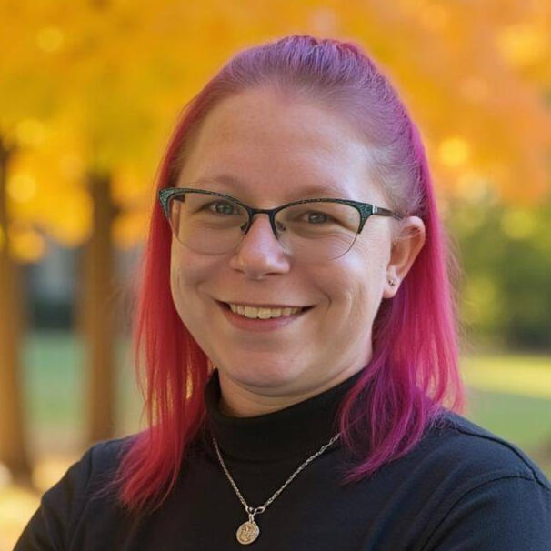 A headshot of Michelle (Hebert) Thomas. Pink hair worn half up half down, green hornrimmed glasses, smiling looking at the camera.